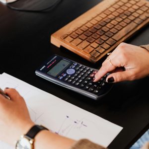 From above crop anonymous female accountant working with calculator and writing down notes on paper while sitting at table with computer keyboard